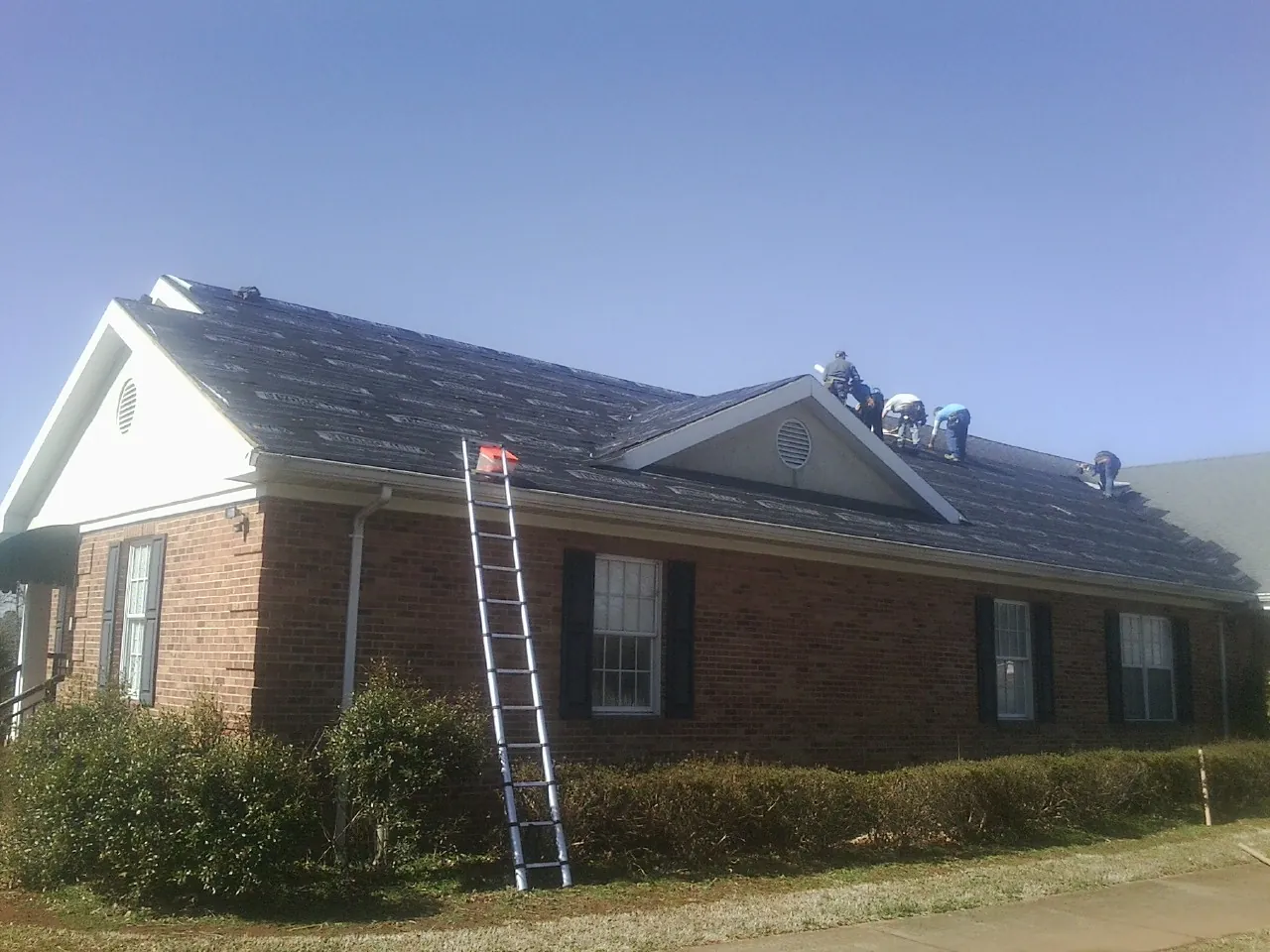 Crew tearing shingles off a brick ranch roof with a ladder set — Emergency Roof Repair work by our crew in Hills